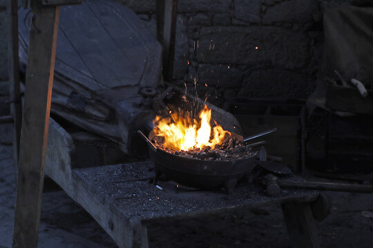 Medieval forge with bright flames and large leather bellows behind the hearth, surrounded by charcoal and traditional blacksmithing equipment in a rustic smithy at a historical fair.