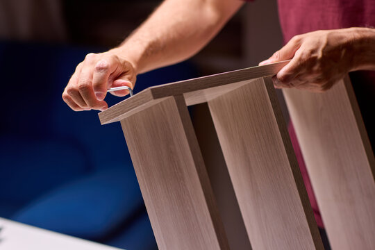 Adult hands cutting stacked corrugated cardboard sheets with utility knife on workbench, close-up of layered edges and precise manual slicing under warm indoor light