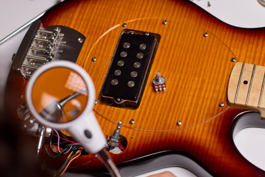 Close up of technician hands repairing electric guitar control cavity under magnifying glass with soldering iron, showing wiring, pickups, screws, and electronic components