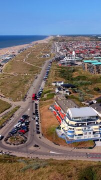 Vertical aerial drone video of coastal boulevard, beachfront hotels, parking roundabout, sand dunes, and North Sea beach coastline in Katwijk aan Zee, South Holland, Netherlands on sunny day