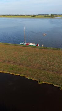Vertical aerial drone video of traditional Dutch sailboat moored at dike embankment, canal waterway, lake, green grass field, and countryside polder landscape in Warmond, South Holland, Netherlands