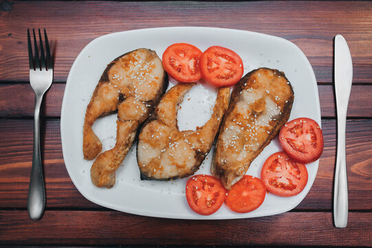 Pieces of fish fried in sesame seeds sit on a white plate next to tomato wedges. Served with a knife and fork on a brown wooden table. A perfect dish for breakfast, lunch, or dinner.