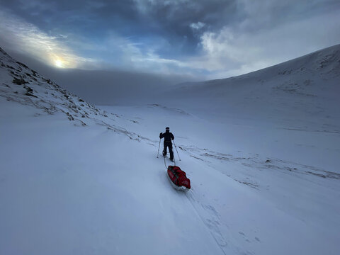 Dovre Municipality, Innlandet, Norway - 18 February 2020: View of a cross-country skier pulling a red sled across the snowy, windswept landscape of Dovrefjell under a dramatic, overcast sky.