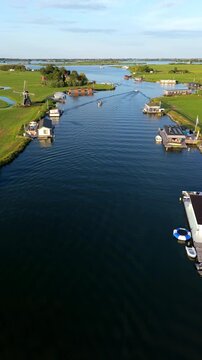Vertical aerial drone video of traditional windmill, modern houseboats, canal waterway, motorboat, and polder landscape in Warmond, South Holland, Netherlands at golden hour