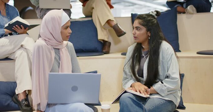 Diverse female classmates planning project on tiered steps after opening laptop, using notebook