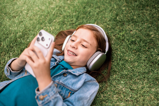 Young child girl lying on the grass listening to music with headphones on mobile phone playlist