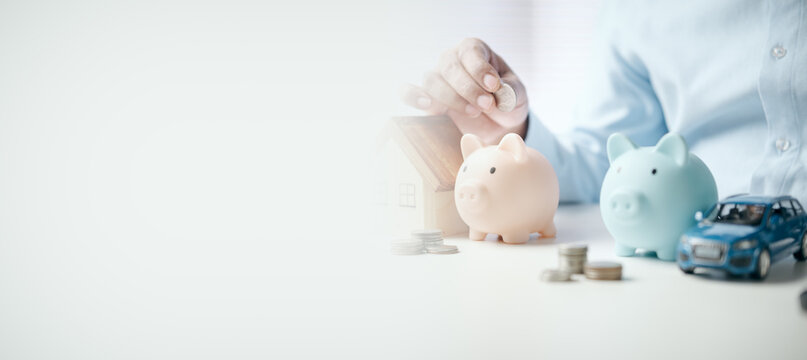 Person saving money with piggy banks, house and car models on white desk with copy space, concept of financial planning, investment, mortgage, and future security for home and vehicle.
