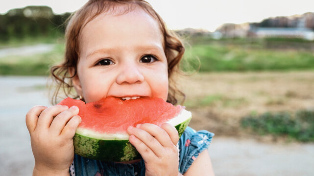 Child toddler eating watermelon in city park during summer vacation - Focus on hands holding fresh fruit - UGC concept