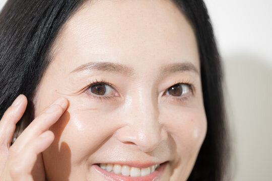 目元のシワをチェックしながら笑顔を見せる日本人ミドル女性、50代のエイジングケアとスキンケアイメージクローズアップ