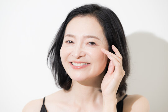 目元に触れながら微笑む日本人ミドル女性、目尻のシワや年齢肌をケアする50代女性のエイジングケアイメージ