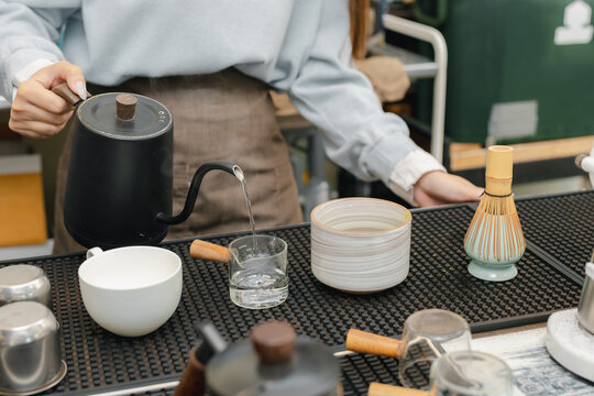 Barista pouring hot water from a gooseneck kettle during matcha tea preparation at a cafe counter with traditional bamboo whisk, ceramic bowl, cup, and professional brewing tools.