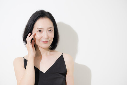 目元に触れながら微笑む日本人ミドル女性、目尻のシワや年齢肌をケアする50代女性のエイジングケアイメージ