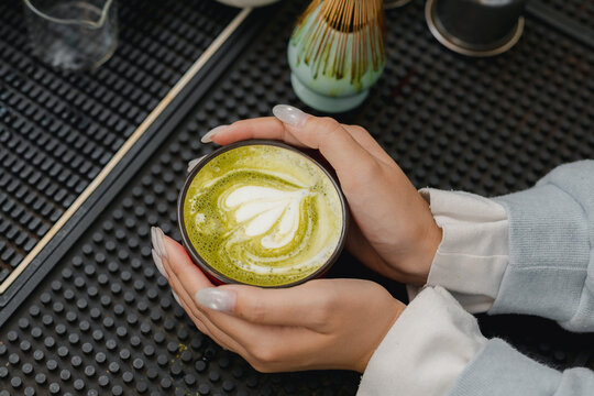 Matcha latte concept. Hands holding fresh green tea drink with creamy latte art at cafe counter, wellness and tea culture. matcha, latte, tea, cafe, drink.