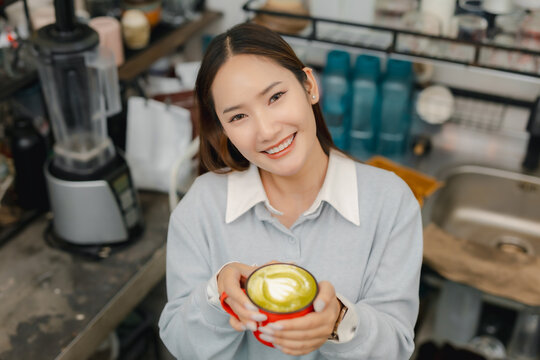 Smiling female barista holding fresh matcha latte with creamy foam art at cafe counter, presenting handcrafted green tea beverage with friendly hospitality and service.