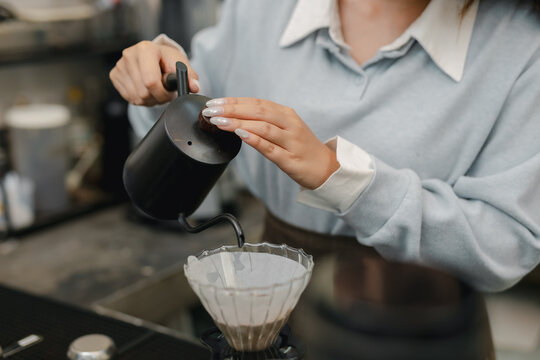 Barista pouring hot water from gooseneck kettle over ground coffee in dripper, preparing fresh pour over coffee with manual brewing technique at cafe counter.