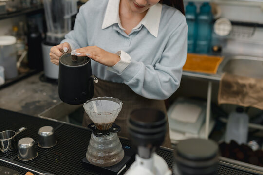 Barista pouring hot water from gooseneck kettle over ground coffee in dripper, preparing fresh pour over coffee with manual brewing technique at cafe counter.