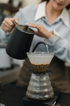 Barista pouring hot water from gooseneck kettle over ground coffee in dripper, preparing fresh pour over coffee with manual brewing technique at cafe counter.