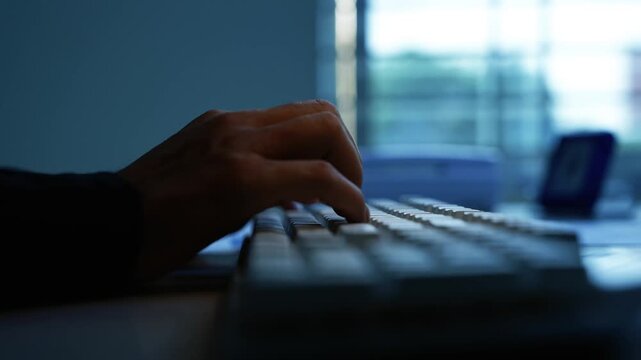 Hand Typing on Keyboard in Dark Blue Office, Close-up of a hand typing on a computer keyboard in a dark room, with blue window light and a blurred laptop screen in the background.