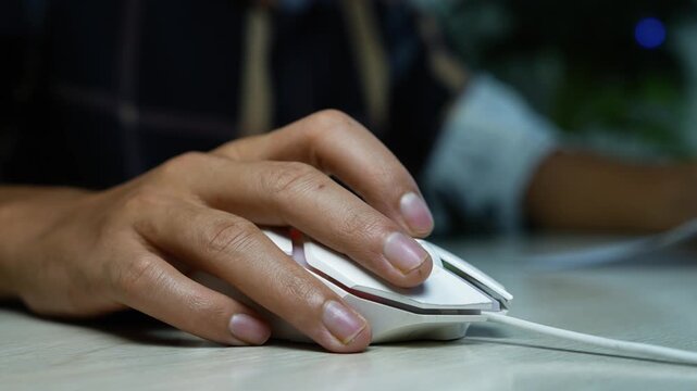 Hand using wired computer mouse on desk, Close-up of a person using a wired computer mouse on a desk, showing online work, browsing, and technology use in a dim office setting.