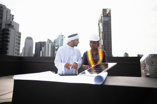 Engineer and investor sizing a photovoltaic addition as the sample panel is angled toward the sun, under harsh tropical daylight on the open roof. Mechanical services in a Bangkok tower.