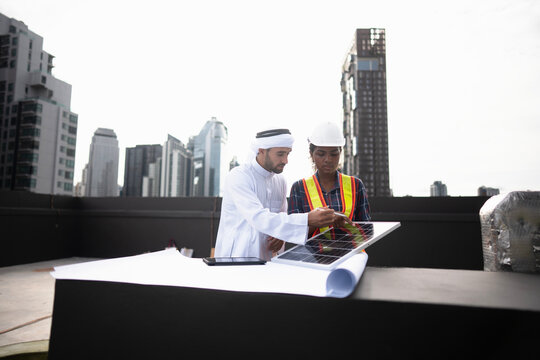 Laptop review of rooftop energy options while the laptop screen is turned toward the client, with the skyline compressed behind the meeting. Planned maintenance on site in Thailand.