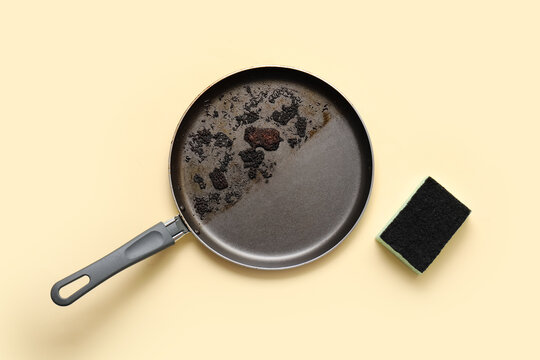 Kitchen cleaning and household chores, dirty burnt frying pan with food residue and abrasive sponge for washing dishes on a light yellow background