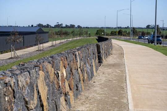 Curved bluestone retaining wall lining a footpath and a nature strip with young trees in a new residential estate, with open fields in Truganina. Landscaped infrastructure in Melbourne, Australia