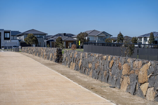 Bluestone stacked rock retaining wall alongside a wide concrete pedestrian footpath in a modern residential estate in Truganina, Melbourne, Victoria, Australia. Stone masonry barriers along sidewalk