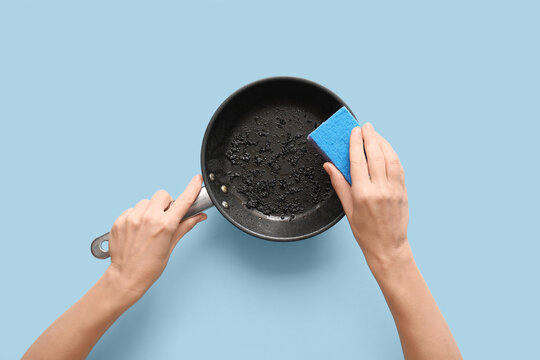 Housework and kitchen cleaning, person scrubbing burnt food residue from a frying pan with a blue sponge on a light blue background