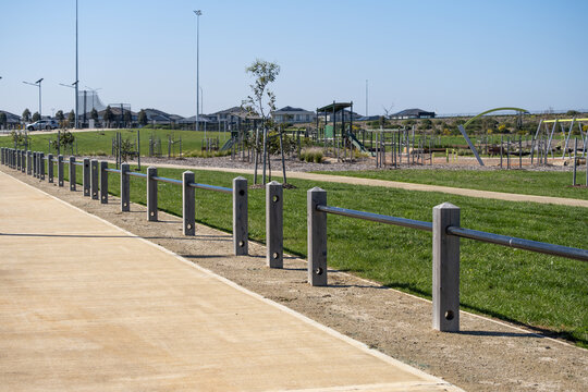 A row of concrete bollards and steel rail fencing runs alongsid a shared path leading to a children's playground in a new residential estate park Truganina, suburban Melbourne, Victoria, Australia.
