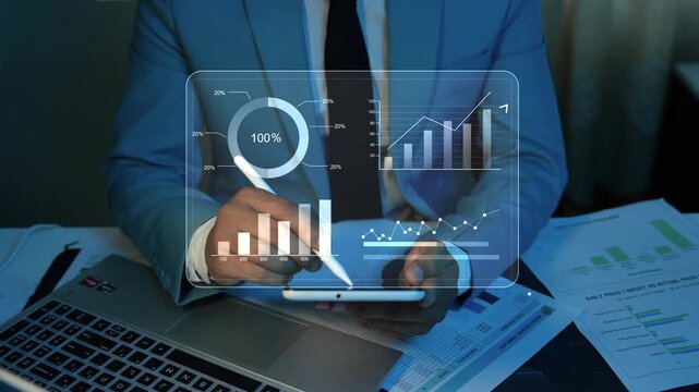 Holographic Financial Charts Animation on th Screen with Businessman Using Tablet, Businessman in a blue suit using a tablet with virtual financial charts, laptop and printed reports on a desk.