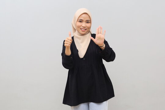 Cheerful, beautiful, smiling Asian girl in hijab, showing six fingers to explain rules as well as steps, standing against white background.