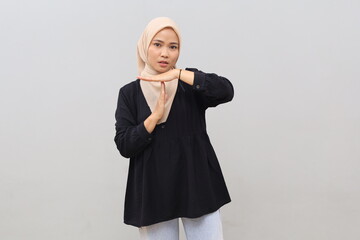 A young Asian woman wearing a headscarf stands against a white wall, making a hand gesture indicating a stop, prohibition, or refusal, while looking at the camera with a serious expression. © ArvStd
