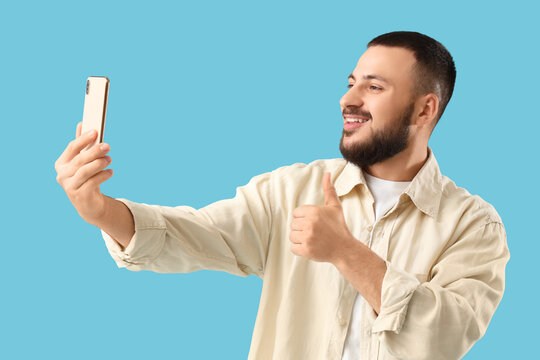 Handsome young man with mobile phone taking selfie and showing thumb-up on blue background