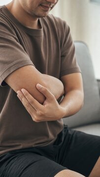 Man in casual t shirt holding aching elbow while sitting on sofa at home interior