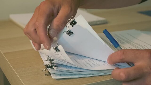 Reviewing Documents and Business Paperwork.Hands reviewing and organizing business documents with a blue pen and binder clips on a desk, representing administration, paperwork, office work, and docume