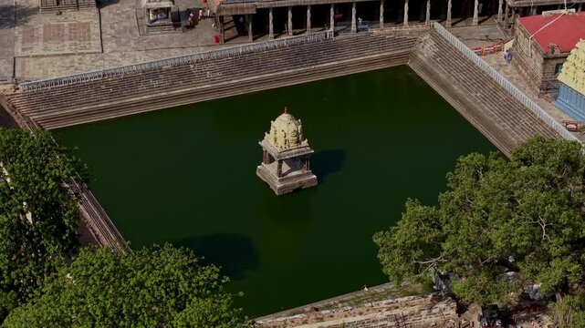 Aerial drone view showing a dark green temple tank with a small shrine at its center. Stone steps surround the water while mature trees frame the historic site in Kanchipuram India