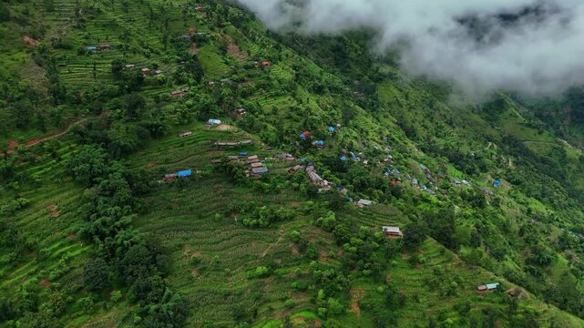 Aerial close up of a rural mountainside village with colorful rooftops nestled among green agricultural terraces as fog rolls over the ridge