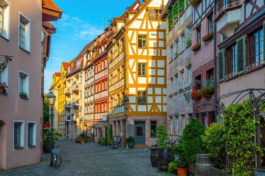 Charming afternoon street view looking down a historic cobblestone alleyway lined with colorful half-timbered buildings, flower boxes, and wine barrels in the old town of Nuremberg