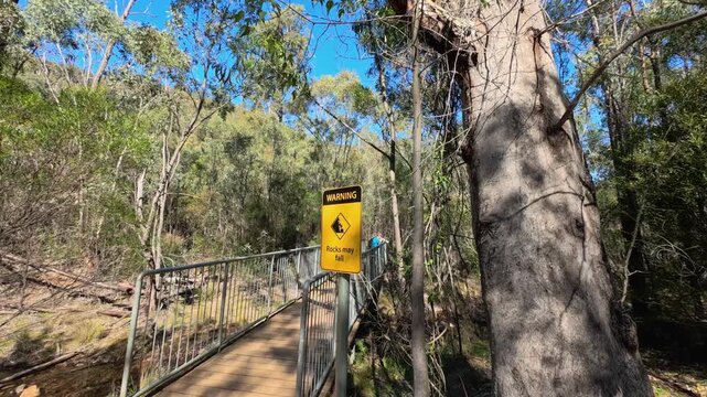 Camera Zooming in on Yellow Rocks May Fall Warning Sign on Forest Footbridge