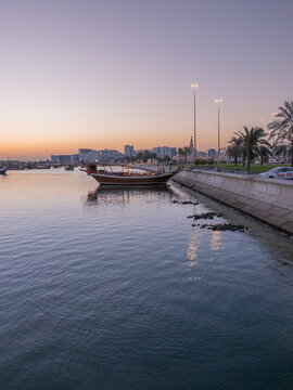 Traditional wooden dhow boat floating along Doha Corniche waterfront promenade with palm trees and city skyline during dusk.