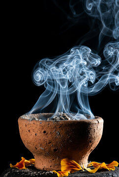 Rustic terracotta incense burner emitting thick, swirling plumes of copal smoke against a dark background, with fresh orange marigold petals arranged on a stone surface at the base.