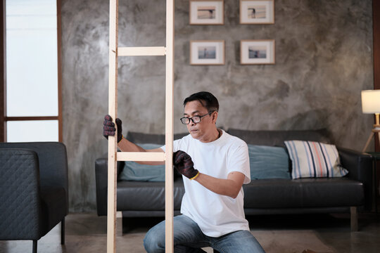 Asian mature man self-assembling flat-pack furniture with DIY tools, checking accuracy and strength of wooden parts, home improvement, craft installation, hobby workshop, handyman active lifestyle.