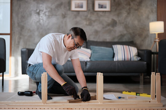 Asian mature man self-assembling wooden flat-pack furniture with DIY carpentry tools, following guide step by step, home improvement, craft installation, hobby workshop, handyman active lifestyle.