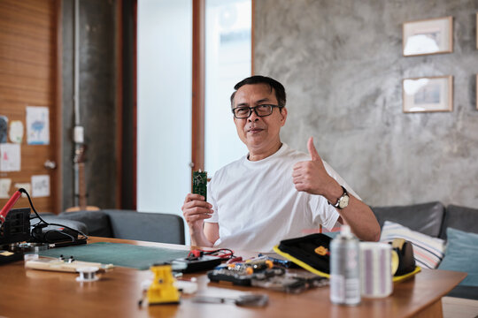 Mature Asian man showing pride in circuit board after successful soldering job, smiling, thumbs up, cheerful job achievement, home electronics appliance repair handyman, DIY fix tools, hobby workshop.