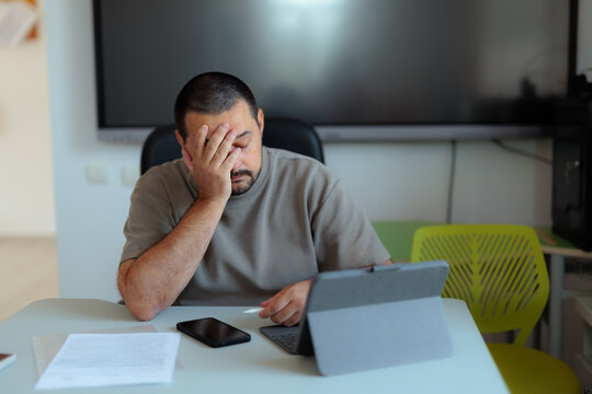 Tired Male Teacher Covering Face With Hand Feeling Stressed In Online Class. Disappointed man looks down covering his face while working on digital tablet