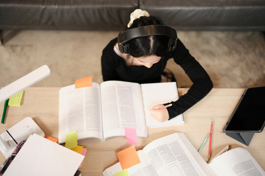 Asian teenage girl in headphones concentrates on learning at home, reading textbooks, note writing, self-tutorial exams, preparation for back to school, Homeschooling, university education lifestyle.
