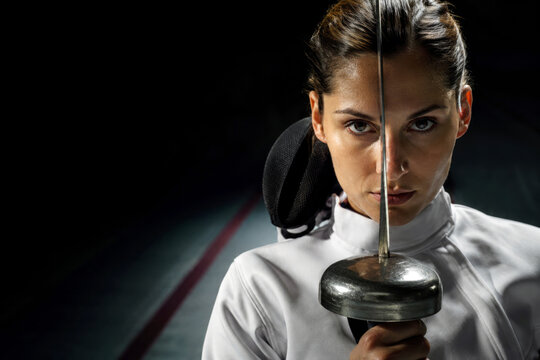 A determined female fencer holds a rapier vertically before her face as a single overhead spotlight reveals her eyes. Intense focus