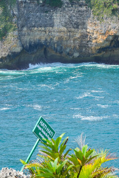 Caution warning sign on a coastal cliff with blue ocean waves crashing against rocky hills in Indonesia.