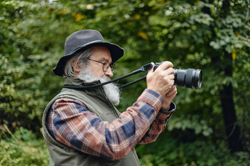Fototapeta premium Caucasian senior man holding digital camera while photographing nature in forest. Image suitable for active retirement concepts, travel marketing, outdoor hobby advertising, side view